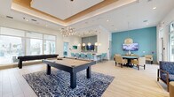 Located at 1061 NE Baker Rd in Stuart, Florida, this single-story community clubhouse showcases a coastal-contemporary design with a cohesive palette of aqua, white, and natural wood tones throughout. The open-concept layout flows seamlessly between multiple functional zones, each thoughtfully appointed for socializing and recreation.

The main gathering area features a coffered ceiling with warm wood trim and ambient cove lighting, anchored by a statement chandelier with organic branching details. A full-size billiards table rests atop a bold navy coral-print area rug, while a shuffleboard table adds additional entertainment options. Light wood-tone flooring runs throughout, enhancing the bright, airy atmosphere.

A built-in kitchenette serves as a central hub, outfitted with a large white quartz island surrounded by turquoise upholstered bar stools, globe pendant lighting, and a light-blue tile accent wall with open wood shelving and stainless steel under-counter refrigerators. An adjacent dining area with woven furniture and a wall-mounted TV provides space for group gatherings.

A separate game nook features a round poker table and wall-mounted television, offering a more intimate setting. The welcoming lounge near the entrance benefits from floor-to-ceiling windows with vibrant blue shades, flooding the space with natural light and views of the surrounding landscape.