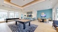Located at 1061 NE Baker Rd in Stuart, Florida, this single-story community clubhouse showcases a coastal-contemporary design with a cohesive palette of aqua, white, and natural wood tones throughout. The open-concept layout flows seamlessly between multiple functional zones, each thoughtfully appointed for socializing and recreation.

The main gathering area features a coffered ceiling with warm wood trim and ambient cove lighting, anchored by a statement chandelier with organic branching details. A full-size billiards table rests atop a bold navy coral-print area rug, while a shuffleboard table adds additional entertainment options. Light wood-tone flooring runs throughout, enhancing the bright, airy atmosphere.

A built-in kitchenette serves as a central hub, outfitted with a large white quartz island surrounded by turquoise upholstered bar stools, globe pendant lighting, and a light-blue tile accent wall with open wood shelving and stainless steel under-counter refrigerators. An adjacent dining area with woven furniture and a wall-mounted TV provides space for group gatherings.

A separate game nook features a round poker table and wall-mounted television, offering a more intimate setting. The welcoming lounge near the entrance benefits from floor-to-ceiling windows with vibrant blue shades, flooding the space with natural light and views of the surrounding landscape.