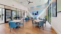 Located at The Reserve in Apopka, Florida, this community gathering space is a well-appointed clubroom designed for socializing and relaxation. The single-floor layout offers a generous open-plan area that feels expansive thanks to soaring double-height ceilings and an abundance of natural light streaming through large, floor-to-ceiling black-framed windows and glass doors that frame views of the landscaped grounds beyond.

The interior design centers on a cohesive navy blue and white color palette, punctuated by warm light wood flooring that runs throughout. A standout feature is the built-in bar counter, finished in white quartz and backed by deep navy cabinetry, set against a striking decorative mosaic tile accent wall with a wall-mounted flat-screen television. Pendant lighting drops elegantly above the bar, where leather counter-height stools invite casual gatherings.

The dining area accommodates multiple tables with modern blue upholstered chairs, providing ample seating for residents. A feature wall with geometric white trim and a soft blue backdrop anchors another mounted television, creating a media-ready lounge zone. Glass partition walls with black steel frames offer a glimpse into adjacent amenity spaces, adding visual depth and connectivity. Ceiling fans, recessed lighting, and curated wall art complete this polished, community-focused space.
