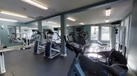 This fitness center is a thoughtfully designed, single-floor workout facility built to support a well-rounded cardio training routine. The space is anchored by a strong selection of cardio equipment, including treadmills, elliptical trainers, and stationary bikes, all arranged to maximize usability and flow throughout the room. Structural columns finished in a muted teal-gray tone divide the space while adding an architectural element that complements the overall color palette of the walls. Large wall mirrors run along one side of the room, visually expanding the space and allowing users to monitor their form during workouts. Multiple windows along the perimeter flood the room with natural light, creating a bright and energizing atmosphere. Overhead fluorescent lighting ensures consistent illumination throughout all hours of use. The dark rubberized flooring provides both durability and safety underfoot, making it well-suited for high-traffic fitness use. Flat-screen monitors are mounted for entertainment or instructional use during workouts. The overall ambiance is clean, functional, and motivating, making this fitness center a practical and inviting space for daily exercise.