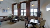 This single-floor space makes a bold architectural statement with its industrial-chic character and refined residential touches. The most commanding features are the towering exposed brick walls and a series of oversized, arched multi-pane windows that stretch dramatically toward the ceiling, bathing the interior in abundant natural light. Roller shades offer flexible light control while preserving the grandeur of the window frames.

The open layout thoughtfully integrates a living area and a dining space. The living area is anchored by a low-profile rectangular coffee table surrounded by patterned upholstered armchairs, creating a comfortable and inviting seating arrangement. A classic floor lamp adds a warm accent to the corner. The dining area features a rich, dark circular table paired with cushioned dining chairs, offering an intimate setting for meals.

Overhead, industrial-style pendant lights hang at varying heights, complementing the raw aesthetic of the brick and the polished nature of the furnishings. The light carpeted flooring provides a soft contrast to the rugged masonry walls. Potted plants and fresh floral arrangements are thoughtfully placed throughout, adding organic warmth and color. The overall ambiance blends historic industrial architecture with comfortable, contemporary living.