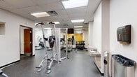 Located at 161 Concord Exchange in South Saint Paul, MN, this fitness center spans a single floor and is divided into two dedicated exercise rooms, offering a well-rounded workout environment. The space features durable rubber flooring throughout, providing a safe and functional surface for a wide variety of exercises.

The main exercise room is generously sized and outfitted with a strong selection of cardio machines, including treadmills and ellipticals, as well as a multi-functional cable machine, strength equipment, a dumbbell rack, and a wall-mounted television — all reflected in full-length mirrors that enhance the sense of space. Accent walls in bold yellow add an energizing visual element, complemented by clean white walls and a drop-panel ceiling with recessed fluorescent lighting that keeps the space bright and well-illuminated.

The secondary exercise room offers a more intimate setting, featuring an inversion table and open floor space ideal for stretching or mat-based workouts. A pass-through window connects the two rooms, maintaining visibility while preserving distinct training zones. A water fountain is conveniently accessible near the main workout area. Together, the two rooms provide a comprehensive and functional fitness facility suitable for a broad range of training needs.