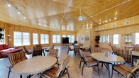 This single-story gathering space exudes a warm, cabin-inspired ambiance through its striking natural pine wood paneling, which lines both the walls and the vaulted ceiling. The architectural highlight is the pitched, coffered wood ceiling, accented by white trim detailing that draws the eye upward and enhances the sense of height and openness throughout the room. Track lighting runs along the ceiling lines, supplemented by a central ceiling fan and light fixture, ensuring the space is well-lit at all times. Large windows wrap around the perimeter of the room, inviting generous amounts of natural light and providing views of the surrounding exterior. The main area is furnished with multiple round tables paired with cushioned, striped armchairs arranged in comfortable conversational clusters, making the space well-suited for social gatherings or community events. A flat-screen display positioned along one wall adds a functional element. Decorative touches, including potted plants and framed artwork, contribute to the inviting atmosphere. The durable, neutral-toned flooring grounds the warm wood tones above, creating a cohesive and welcoming environment throughout.