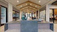 Located at 275 Pine Terrace Dr in Conroe, TX, this single-floor community clubhouse offers a generously sized, thoughtfully designed gathering space that makes a striking first impression. At the heart of the space is a dramatic vaulted ceiling with exposed wood beams running its full length, complemented by recessed lighting and elegant pendant chandeliers that cast a warm, inviting glow throughout.

Guests are welcomed at a sleek reception desk adorned with decorative teal ceramic vases, positioned at the entrance to orient visitors into the expansive interior beyond. The open floor plan flows seamlessly from the reception area into multiple distinct lounge zones, each furnished with curated seating arrangements including upholstered armchairs, sectionals, and accent tables set atop patterned area rugs.

Floor-to-ceiling windows along the perimeter flood the space with abundant natural light and provide clear views to the surrounding exterior. A dedicated entertainment area features a bold geometric accent wall housing a flat-screen television and a billiards table, while a bar-height counter with gold-accented stools adds a social focal point. A separate workstation area with built-in desks and modern task chairs offers a functional workspace. Large-format light tile flooring unifies the entire layout, enhancing the sense of openness and scale throughout this impressive amenity space.