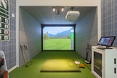 Located in San Jose, this dedicated indoor golf simulator room is a meticulously designed space that delivers an immersive golfing experience. The room is finished with lush artificial turf underfoot, marked with white yardage lines that reinforce the course atmosphere from the moment you step inside.

The centerpiece of the space is a large-format HD Golf simulation screen, ceiling-mounted projector, and a designated hitting mat, allowing players to tee off on virtual courses with realistic visuals of rolling fairways and mountain backdrops. Track lighting overhead ensures the hitting zone is well-illuminated for optimal play.

One wall is covered in bold plaid wallpaper and serves as the backdrop for a prominent US Open-style scoreboard display, adding a tournament-worthy aesthetic. The motivational phrase "Drive for show, Putt for dough!!" is emblazoned in red lettering on the upper white wall, reinforcing the playful yet passionate theme throughout.

A golf bag storage rack along the feature wall keeps equipment neatly organized, while a pair of plush navy blue lounge chairs provides comfortable seating for spectators and players awaiting their turn. The soaring ceiling height amplifies the sense of openness within the room. A glass-paneled entry door allows easy access and connects the simulator bay to the surrounding facility.