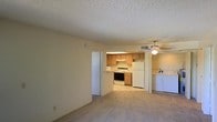 This single-floor unit offers an open layout that seamlessly connects the living area to the kitchen, creating a practical and cohesive living space. The living area is finished with wall-to-wall carpeting underfoot and features a ceiling fan with a light fixture, contributing to a comfortable, well-ventilated atmosphere. The textured popcorn ceiling runs throughout, adding a classic residential touch.

The kitchen is visible from the main living area and is equipped with medium-toned wood cabinetry providing ample storage. White appliances, including a freestanding electric range, a microwave, and a full-sized refrigerator, are all included. The kitchen's tile or linoleum flooring contrasts with the carpeted living space, delineating the two areas naturally.

A doorway adjacent to the kitchen leads to a dedicated laundry area, where a side-by-side washer and dryer are housed — a convenient in-unit feature. A separate doorway near the living area provides access to additional rooms within the unit. Overall, the space offers a straightforward, functional layout suited for everyday living.