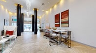 This stunning lobby entrance is defined by its soaring double-height ceilings and an expansive, open layout that immediately conveys a sense of grandeur. The floors throughout are finished in a light-toned wood-look herringbone pattern, adding warmth and visual texture to the space. Tall, bold black cylindrical columns rise dramatically from floor to ceiling, creating a striking architectural contrast against the crisp white walls. A cascading crystal chandelier with gold ceiling medallions serves as a focal centerpiece, bathing the space in ambient light alongside recessed and cove lighting that accentuates the ceiling's height.

The main lobby area is divided into distinct functional zones. A lounge seating area features a neutral-toned sofa, high-back accent chairs with bold red and copper pillows, glass-framed coffee tables, and a curated gallery wall of six geometric framed artworks. A communal bar-height table with upholstered stools lines one wall beneath a pair of large, vibrant red abstract paintings.

The entrance vestibule is elegantly composed with symmetrical high-back white wing chairs, built-in niche displays, sheer curtained windows, and frosted glass entry doors. Throughout, the design balances modern sophistication with residential warmth, reflecting a polished, upscale community aesthetic managed by Madera Residential.