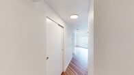 This single-floor residence offers a well-organized layout with a clean, contemporary aesthetic defined by crisp white walls, hardwood-style flooring throughout the main living areas, and an abundance of natural light. The open-concept kitchen and living area forms the heart of the unit, where a peninsula counter creates a natural divide between the two spaces. The kitchen is outfitted with stainless steel appliances — including a refrigerator, range, and built-in oven — complemented by white cabinetry, neutral countertops, and a double sink beneath a window. A ceiling fan with integrated lighting anchors the living area, which receives generous natural light through multiple windows. A hallway connects the main living space to the private areas of the unit, with a sliding-door closet providing additional storage along the corridor. The bedroom is finished with plush carpeting, a ceiling fan, a baseboard heater, and a window that brings in natural light. A dedicated walk-in closet off the bedroom offers ample storage with a built-in hanging rod. The bathroom features hardwood-style flooring and includes a soaking tub. Throughout the unit, the design emphasizes simplicity, functionality, and a bright, welcoming atmosphere.
