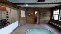 This indoor wellness area offers a self-contained retreat designed for relaxation and recreation. The space is defined by its warm, rustic aesthetic, with wood-paneled walls that create a cozy, lodge-like ambiance throughout. The centerpiece of the room is a built-in hot tub, finished with brick tile surround and textured flooring that provides both comfort and safety underfoot. Adjacent to the hot tub area, a dedicated sauna room is accessible through a wooden door, offering an additional layer of wellness amenity within the same space. The ceiling features recessed lighting that provides soft, functional illumination, complementing the enclosed and intimate atmosphere. A large sliding glass door or window on one side of the room allows for ventilation and a connection to an adjacent area. Posted signage outlines spa rules and safety guidelines, noting that the hot tub is restricted to guests 14 years of age and older. The overall design prioritizes function and comfort, making this space a practical and inviting wellness facility suited for year-round use.