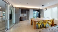 Located at 401 Mt Vernon St in Boston's Dorchester neighborhood, this single-floor space is designed with a modern, contemporary aesthetic that balances comfort and functionality. The lounge area serves as a central gathering point, anchored by a bold deep-teal geometric accent wall adorned with a wall-mounted flat-screen TV. Seating options include a leather sectional sofa, vibrant chartreuse pod chair, and rounded teal and navy accent chairs, all arranged atop a striking chevron-patterned area rug over light-toned flooring. Natural light filters in through windows fitted with roller shades, contributing to the bright and welcoming ambiance. A glass-partitioned entryway with sleek aluminum framing connects the space to adjacent areas, creating an open and transparent flow. The kitchenette area is thoughtfully outfitted with dark wood cabinetry, stainless steel appliances including a full-size refrigerator, and a sink, complemented by vending machines for added convenience. A solid wood communal dining table surrounded by yellow metal chairs provides an ideal setting for casual dining or collaboration. Recessed lighting and cove lighting throughout enhance the polished, inviting atmosphere of this well-appointed space.