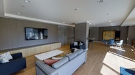 This single-floor space is designed as a modern, open-plan communal area that seamlessly integrates multiple functional zones. The main lounge area is anchored by a large wall-mounted flat-screen television set against a sleek built-in media console with light wood cabinetry, providing ample storage while maintaining a clean aesthetic. Comfortable gray upholstered sofas and accent chairs are arranged around a minimalist white coffee table, creating an inviting seating area well-suited for relaxation or social gatherings.

Warm hardwood-style flooring runs throughout the space, contributing to a cohesive and contemporary feel. Neutral gray walls are complemented by recessed ceiling lighting, ensuring the area is well-lit. Vertical sheer curtains along one wall soften the space and allow for diffused natural light.

Adjacent to the main lounge, a secondary seating area features additional chairs and a raised countertop with bar-height seating, functioning as a casual dining or social space. A structural column subtly divides the zones while maintaining an open, airy atmosphere. The overall design balances functionality and modern aesthetics, making it an well-appointed shared living space.
