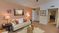 This single-floor space presents a thoughtfully designed living area anchored by warm terracotta and neutral tones that create an inviting, cozy atmosphere. The living room serves as the focal point of the space, featuring plush, beige wall-to-wall carpeting that adds softness underfoot. A textured ceiling with a ceiling fan enhances the room's character while providing practical airflow.

The accent wall, painted in a rich salmon-peach hue, draws immediate attention and provides a warm backdrop for a large, ornate mosaic-framed mirror that reflects light throughout the room. Decorative wall art, including a lattice-style wooden piece, adds dimension and visual interest to the neutral-toned walls.

The living area comfortably accommodates a cream-colored sofa adorned with copper and rust-toned accent pillows, complemented by rustic wooden side tables and a cowhide-style area rug that grounds the seating arrangement. A floor lamp with a warm-toned shade provides ambient lighting, enhancing the room's welcoming character.

A hallway visible from the living area leads to additional rooms, suggesting a practical and well-connected layout. The overall design balances warmth, texture, and decorative detail to create a cohesive and comfortable living environment.