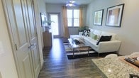 This well-appointed single-floor residence offers a thoughtfully designed interior that balances modern style with everyday comfort. The living area serves as the heart of the space, showcasing rich, dark hardwood-style flooring that extends seamlessly throughout the main areas, creating a warm and cohesive aesthetic. A white sectional sofa with decorative accent pillows anchors the room, complemented by a sleek glass and chrome coffee table and a bold striped area rug that adds visual interest underfoot. A ceiling fan with integrated lighting provides both comfort and ambiance overhead. Natural light pours in through multiple windows dressed with sheer panels and warm-toned drapes, giving the space a bright and inviting atmosphere. A glass door at the far end of the living area hints at outdoor access, further enhancing the sense of openness. Framed wall art and lush green plants add personality and a touch of nature to the interior. Adjacent to the living area, a granite countertop surface suggests a well-equipped kitchen with quality finishes. The overall design aesthetic is clean, contemporary, and carefully curated, making this residence a comfortable and visually appealing living environment.