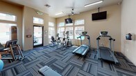 This single-floor fitness center is thoughtfully designed to support a well-rounded workout experience. The space is anchored by a selection of cardio equipment, including treadmills, an elliptical, and a recumbent bike, all arranged to allow comfortable use and clear sightlines. Multiple wall-mounted televisions are positioned throughout the room for entertainment during workouts.

A dedicated strength and stretching area features a dumbbell rack with a full range of free weights, an adjustable weight bench, and various exercise accessories such as stability balls and resistance bands. The flooring consists of durable, patterned carpet tiles in a blue-grey geometric design that adds visual depth to the space.

Natural light fills the room through a generous arrangement of windows, offering views of an outdoor pool and surrounding buildings. A bold yellow accent wall displays an array of motivational quotes from notable figures, creating an energizing and encouraging atmosphere.

The entrance features a glass door with a branded welcome mat, while a ceiling fan and overhead lighting ensure the space remains comfortable year-round. The overall design balances functionality with an uplifting aesthetic, making it a practical and motivating environment for residents.