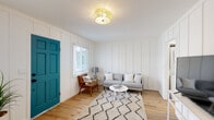 This thoughtfully designed single-floor space blends coastal charm with modern comfort through a cohesive white-and-teal color palette. The living area is defined by striking board-and-batten wall paneling painted crisp white, warm-toned hardwood floors, and a bold teal entry door that adds a vibrant pop of color. A gold drum flush-mount ceiling fixture provides warm ambient lighting, complementing the natural light that filters through white-blinded windows. The living room comfortably accommodates a grey sofa, a mid-century leather accent chair, a round white coffee table, and a plush geometric-patterned area rug, creating an inviting and well-appointed gathering space. A dedicated nook features a mini-split HVAC unit, natural greenery, and artful décor, lending a serene, meditative quality to the layout. The galley-style kitchen is a true standout, showcasing retro turquoise appliances — including a gas range with matching hood and a full-size refrigerator — set against white shaker cabinetry and light marble-look tile backsplash. The bathroom is elegantly finished with glossy subway tile surrounding the tub and shower, a vintage-style bridge faucet, and a classic white vanity. In-unit laundry with a stacked washer and dryer rounds out this efficient, stylish space.