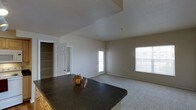 This single-story space showcases an open-concept design that seamlessly connects the kitchen and living areas. The kitchen features a dark granite-topped island with warm wood cabinetry, providing both prep space and a natural visual anchor for the layout. The countertop's reflective surface adds a polished, functional element to the space.

The living area is finished with neutral-toned carpet, creating a comfortable and cohesive atmosphere throughout. Vaulted ceilings with a textured finish add a sense of vertical dimension, enhancing the overall feeling of openness. The walls are painted in a soft, neutral tone that complements the light flooring and contributes to a clean, understated aesthetic.

Natural light is a prominent feature, with a large window fitted with horizontal blinds on one side and a glass-paneled exterior door introducing additional brightness and providing access to the outdoors. A doorway visible from the main area leads to additional spaces, including what appears to be a closet with built-in shelving, suggesting practical storage solutions throughout the unit. The overall ambiance is bright, airy, and move-in ready.