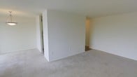 This single-floor space presents a straightforward and functional layout designed to maximize usability. The main living area is finished with wall-to-wall carpeting in a neutral beige tone, providing a soft underfoot feel that complements the clean, white walls throughout. A prominent partial wall divides the space, creating a natural separation between areas while maintaining an open and airy feel. This architectural element adds subtle definition to the layout without fully enclosing individual zones. To the left of the dividing wall, a glimpse into an adjacent area suggests additional functional space, possibly a utility or kitchen area, identifiable by what appears to be cabinetry or shelving visible in the background. On the right side of the dividing wall, a hallway leads further into the unit, hinting at additional rooms beyond the main living space. Electrical outlets are positioned at standard heights along the baseboards, offering practical placement for furnishings. The white walls and light-toned carpet create a bright, neutral palette throughout, making the space highly adaptable to a variety of interior styles and configurations. Overall, the space conveys a clean, move-in-ready environment.