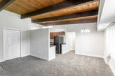 This single-floor residence showcases a distinctive architectural character defined by its prominent dark wood beams and warm tongue-and-groove wood plank ceilings, which lend a rustic yet refined ambiance throughout the main living areas. The open-concept living space is generously proportioned, finished with neutral carpet flooring and light gray walls that create a bright, welcoming atmosphere. A semi-open partition wall separates the living area from the kitchen, maintaining an airy flow between the two spaces. The kitchen is equipped with rich dark walnut cabinetry, dark granite-style countertops, a stainless steel sink, a black refrigerator, and a dishwasher, offering both style and practicality. Pendant lighting and a chandelier add tasteful illumination throughout the main living area. The bedrooms continue the wood-beamed ceiling aesthetic and are carpeted, with one bedroom featuring a wide horizontal window that brings in natural light and outdoor views. A spacious walk-in closet with full wire shelving and hanging rods provides ample storage. The bathroom includes a combined tub and shower unit with white tile surround, complemented by wood-look flooring. A sliding glass door in the living area provides access to an outdoor space, further enhancing the unit's connection to natural light.