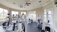 This fitness center is designed to support a comprehensive workout experience within a bright and open single-floor layout. The space is outfitted with a wide selection of strength training machines, including cable and pulley systems, weight machines, and a stationary bike, alongside cardio equipment visible along the perimeter. The dark rubber flooring provides both safety and durability underfoot, while the neutral-toned walls contribute to a clean, focused environment. Multiple large windows line the walls, offering views of the surrounding outdoor greenery and allowing abundant natural light to fill the room throughout the day. Crown molding adds a refined architectural detail along the upper walls, lending a polished feel to the functional space. Overhead, several ceiling fans are evenly distributed across the room, ensuring consistent air circulation during workouts. Recessed lighting supplements natural light for use during all hours. The layout is thoughtfully arranged to allow users to move freely between equipment stations, making the space practical for individual training sessions. Overall, the fitness center balances functionality with a comfortable and well-maintained atmosphere.