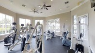 This fitness center is designed to support a comprehensive workout experience within a bright and open single-floor layout. The space is outfitted with a wide selection of strength training machines, including cable and pulley systems, weight machines, and a stationary bike, alongside cardio equipment visible along the perimeter. The dark rubber flooring provides both safety and durability underfoot, while the neutral-toned walls contribute to a clean, focused environment. Multiple large windows line the walls, offering views of the surrounding outdoor greenery and allowing abundant natural light to fill the room throughout the day. Crown molding adds a refined architectural detail along the upper walls, lending a polished feel to the functional space. Overhead, several ceiling fans are evenly distributed across the room, ensuring consistent air circulation during workouts. Recessed lighting supplements natural light for use during all hours. The layout is thoughtfully arranged to allow users to move freely between equipment stations, making the space practical for individual training sessions. Overall, the fitness center balances functionality with a comfortable and well-maintained atmosphere.