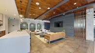 Located at 1170 Temple Dr in Sugar Hill, Georgia, this single-floor amenity space showcases a sophisticated blend of contemporary design and industrial-inspired architecture. The main lounge area is anchored by rich wood plank ceilings with exposed dark steel beams, large-format stone tile flooring, and a dramatic slate-tiled feature wall housing a wall-mounted display screen. Comfortable seating arrangements — including a caramel leather sofa and white upholstered chairs — create inviting gathering zones throughout the open layout. A standout design element is the row of arched, teal-tiled nooks with built-in bench seating and sculptural wall sconces, adding a boutique-hotel ambiance. The kitchen area features warm wood cabinetry, white quartz countertops, open iron-framed shelving, a zellige tile backsplash, and stainless steel appliances, complemented by patterned mosaic floor tile accents. A cozy windowed nook with plaid banquette seating and views to the exterior courtyard offers a more intimate setting. An industrial-style mezzanine corridor with an expanded metal mesh railing and exposed concrete ceiling connects the spaces, while a bright entry vestibule with floor-to-ceiling glass doors and large-format marble tile flooring provides a welcoming arrival experience with outdoor courtyard views.