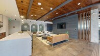 Located at 1170 Temple Dr in Sugar Hill, Georgia, this single-floor amenity space showcases a sophisticated blend of contemporary design and industrial-inspired architecture. The main lounge area is anchored by rich wood plank ceilings with exposed dark steel beams, large-format stone tile flooring, and a dramatic slate-tiled feature wall housing a wall-mounted display screen. Comfortable seating arrangements — including a caramel leather sofa and white upholstered chairs — create inviting gathering zones throughout the open layout. A standout design element is the row of arched, teal-tiled nooks with built-in bench seating and sculptural wall sconces, adding a boutique-hotel ambiance. The kitchen area features warm wood cabinetry, white quartz countertops, open iron-framed shelving, a zellige tile backsplash, and stainless steel appliances, complemented by patterned mosaic floor tile accents. A cozy windowed nook with plaid banquette seating and views to the exterior courtyard offers a more intimate setting. An industrial-style mezzanine corridor with an expanded metal mesh railing and exposed concrete ceiling connects the spaces, while a bright entry vestibule with floor-to-ceiling glass doors and large-format marble tile flooring provides a welcoming arrival experience with outdoor courtyard views.