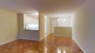 This single-floor space showcases an open-concept layout that promotes a seamless flow between living and functional areas. The warm, honey-toned parquet flooring runs throughout, adding a classic character and a polished finish underfoot.

The kitchen is positioned to one side and features white cabinetry with under-cabinet lighting, a pass-through window opening, and a sink along the counter — allowing visual connectivity to the main living area. Adjacent to the kitchen, a built-in peninsula counter with white upper cabinetry and a two-tone lower section serves as a natural dividing element between zones without fully closing off the space.

Two pendant lights hang from the ceiling, casting warm, focused illumination that enhances the airy feel of the open layout. The neutral beige and white wall tones complement the warm flooring and contribute to a bright, cohesive atmosphere throughout.

A hallway visible toward the rear leads to additional areas of the unit, with a white door suggesting access to a separate room or entryway. The overall design is clean, understated, and functional, making efficient use of the available floor space.