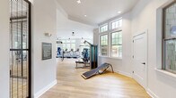 Located at 1421 Legacy Falls Dr in Wake Forest, NC, this comprehensive fitness facility spans a single floor and offers an impressive array of amenities designed to support a wide range of physical activities. The space is organized into two distinct exercise rooms connected by a hallway, each serving a different fitness purpose.

The main exercise room is a bright, open area bathed in natural light from large multi-pane windows along the perimeter. Light hardwood flooring runs throughout, complementing the clean white walls and tray ceilings fitted with pendant and ceiling fan fixtures. This room is outfitted with a full row of treadmills, ellipticals, a multi-function cable machine, TRX suspension trainers, a heavy punching bag, free weights, and benches—providing a well-rounded cardio and strength training environment.

A second exercise room features a striking dark-painted ceiling that adds a bold, focused atmosphere. It is equipped with spin bikes, kettlebells, stability balls, resistance tools, and full-length mirrors, making it ideal for group fitness or functional training sessions.

The facility also includes a soaring indoor basketball court with a regulation backboard and red court markings on polished hardwood floors, enclosed by tall industrial-style metal grid partitions. High clerestory windows flood the court with natural light. A dedicated free weights area with squat racks, benches, and mirrored walls rounds out the offerings.