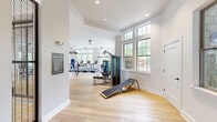 Located at 1421 Legacy Falls Dr in Wake Forest, NC, this comprehensive fitness facility spans a single floor and offers an impressive array of amenities designed to support a wide range of physical activities. The space is organized into two distinct exercise rooms connected by a hallway, each serving a different fitness purpose.

The main exercise room is a bright, open area bathed in natural light from large multi-pane windows along the perimeter. Light hardwood flooring runs throughout, complementing the clean white walls and tray ceilings fitted with pendant and ceiling fan fixtures. This room is outfitted with a full row of treadmills, ellipticals, a multi-function cable machine, TRX suspension trainers, a heavy punching bag, free weights, and benches—providing a well-rounded cardio and strength training environment.

A second exercise room features a striking dark-painted ceiling that adds a bold, focused atmosphere. It is equipped with spin bikes, kettlebells, stability balls, resistance tools, and full-length mirrors, making it ideal for group fitness or functional training sessions.

The facility also includes a soaring indoor basketball court with a regulation backboard and red court markings on polished hardwood floors, enclosed by tall industrial-style metal grid partitions. High clerestory windows flood the court with natural light. A dedicated free weights area with squat racks, benches, and mirrored walls rounds out the offerings.
