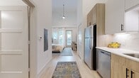 This efficiently designed 1-bedroom, 1-bathroom unit offers a thoughtful layout that maximizes every square foot. The open entryway leads directly into a compact kitchen equipped with stainless steel appliances — including a full-size refrigerator and dishwasher — warm wood cabinetry, light gray upper cabinets, a marble-style tile backsplash, and a single-basin sink. Light wood-tone flooring runs throughout the entire unit, lending a cohesive and airy feel.

The generously proportioned bedroom is a standout feature, boasting soaring ceilings that amplify the sense of space. A modern pendant ceiling fan provides both style and function. Large windows and a glass-paned exterior door flood the room with natural light and offer direct outdoor access. A dedicated closet provides practical storage.

The bathroom is clean and contemporary, featuring a tub-shower combination with shiplap-style white tile surround, a wood-toned vanity with white countertop, black hardware accents, and overhead cabinetry for added storage.

Throughout the unit, crisp white walls, high ceilings, and consistent flooring create a bright, modern ambiance that feels open and well-appointed despite its compact footprint.
