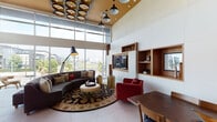 Located on Chandler Boulevard in Los Angeles, this single-floor space is a thoughtfully designed communal lounge that blends contemporary architecture with carefully curated interior details. The space is defined by dramatically tall ceilings that create an open, airy atmosphere, complemented by expansive floor-to-ceiling windows that flood the interior with natural light and offer views of the surrounding North Hollywood neighborhood, including palm trees and adjacent urban streetscapes.

The lounge area features a curved sectional sofa anchored by an ornate area rug and a round upholstered ottoman, accompanied by vibrant red accent chairs and a built-in wall unit of warm wood shelving housing media and display space. An articulating floor lamp adds a designer touch to this seating arrangement. The ceiling above is distinguished by a warm wood panel inset adorned with circular, layered acoustic or decorative elements, adding visual texture overhead.

A wet bar area showcases herringbone-patterned tile in warm metallic tones, wood cabinetry with dark countertops, and a series of globe pendant lights in brass. A shuffleboard table nearby offers a recreational focal point. A separate dining nook features a solid wood table with matching chairs beneath a trio of framed art prints, completing the space with a refined, welcoming ambiance.