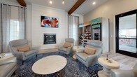 Located on Carl D Silver Pkwy in Fredericksburg, Virginia, this single-floor community clubhouse offers a well-appointed gathering space designed for both social and recreational use. The grand entrance lobby immediately impresses with soaring ceilings, exposed wood ceiling beams, and distinctive large-format hexagonal tile flooring. A substantial round pedestal table anchors the center of the entryway, flanked by a long white bar-height counter lined with velvet blue barstools. Bold artwork and carefully chosen furnishings contribute to a polished yet welcoming atmosphere, while glass doors open directly to an outdoor pool area, flooding the space with natural light.

The interior flows into multiple functional zones. A game lounge features a shuffleboard table, wall-mounted flat-screen TVs, and vaulted ceilings with rustic wood beams set against teal-toned walls. An adjoining kitchen area showcases dark navy cabinetry with white countertops, exposed beam ceilings, and ample counter space. A dedicated dining area provides high-top communal tables paired with gold-toned bistro chairs beneath large windows with layered drapery. A cozy lounge nook includes a fireplace, comfortable seating, and a retail refresh station for added convenience.