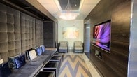 This single-floor space showcases a sophisticated contemporary design that seamlessly blends comfort and style. The main living area is anchored by a plush, tufted gray sectional sofa adorned with accent pillows, complemented by a set of sleek ottomans that double as additional seating or surface space. A striking chevron-patterned rug in gray and white tones defines the lounge area, adding a bold geometric element to the floor.

Two accent chairs with textured upholstery are positioned opposite the sofa, creating an intimate and balanced seating arrangement. A large, wall-mounted flat-screen television is set into a dark wood panel feature wall, adding both a functional and architectural focal point to the room.

Overhead, a cascading crystal chandelier serves as a glamorous centerpiece, drawing the eye upward toward a recessed coffered ceiling detail that adds dimension and depth to the space. The walls are finished in a light neutral tone, providing a clean backdrop that highlights a vibrant abstract artwork hung prominently in the room.

The overall ambiance is polished and modern, with dark wood accents, layered lighting, and curated furnishings working together to create a space that feels both luxurious and inviting.
