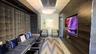 This single-floor space showcases a sophisticated contemporary design that seamlessly blends comfort and style. The main living area is anchored by a plush, tufted gray sectional sofa adorned with accent pillows, complemented by a set of sleek ottomans that double as additional seating or surface space. A striking chevron-patterned rug in gray and white tones defines the lounge area, adding a bold geometric element to the floor.

Two accent chairs with textured upholstery are positioned opposite the sofa, creating an intimate and balanced seating arrangement. A large, wall-mounted flat-screen television is set into a dark wood panel feature wall, adding both a functional and architectural focal point to the room.

Overhead, a cascading crystal chandelier serves as a glamorous centerpiece, drawing the eye upward toward a recessed coffered ceiling detail that adds dimension and depth to the space. The walls are finished in a light neutral tone, providing a clean backdrop that highlights a vibrant abstract artwork hung prominently in the room.

The overall ambiance is polished and modern, with dark wood accents, layered lighting, and curated furnishings working together to create a space that feels both luxurious and inviting.