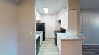 This single-floor unit offers a functional and well-appointed layout designed for comfortable everyday living. The open living and dining area serves as the heart of the space, featuring durable wood-style flooring, neutral beige walls, and a textured ceiling. A ceiling fan with a light fixture provides both comfort and illumination, while vertical blinds on the window allow for adjustable natural light. The entry door opens directly into this main living area, creating a straightforward and accessible flow throughout.

The galley-style kitchen is efficiently designed, lined on both sides with crisp white cabinetry and granite-patterned countertops. Black appliances — including a refrigerator, electric range, and dishwasher — offer a clean contrast to the bright cabinetry. Light wood-style flooring and a skylight-style overhead fixture keep the kitchen bright and practical.

The bedroom is a quiet, carpeted retreat with warm neutral tones, a window fitted with horizontal blinds for privacy, and a ceiling-mounted light fixture. The layout from the 3D model also indicates a bathroom accessible from the main hallway, with additional storage and utility features integrated into the overall floor plan. The space presents a clean, move-in-ready aesthetic throughout.