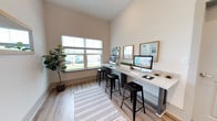 Located at 300 Bellamy Ave in Conway, South Carolina, this single-floor space is designed to serve as a practical and welcoming community hub. The interior showcases a well-appointed computer lounge area that combines functionality with a clean, modern aesthetic. A built-in granite countertop workstation runs along the wall, accommodating multiple desktop computers and providing ample workspace for residents or visitors. Sleek black metal bar stools offer comfortable seating at the stations. Natural light pours in through large windows fitted with horizontal blinds, creating a bright and open atmosphere while offering views of the surrounding greenery and parking area. The walls are finished in a warm neutral tone, complemented by wood-look plank flooring and a patterned area rug that adds a touch of softness underfoot. Framed whiteboards and display panels are mounted along the wall, serving as informational and organizational tools. Subtle greenery accents the space with small potted plants placed along the countertop. The high ceilings contribute to an airy, spacious feel, making the area conducive to focused work or casual browsing in a comfortable, community-oriented environment.
