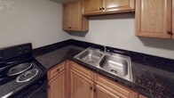 This single-floor unit offers a straightforward, functional layout unified by light beige ceramic tile flooring throughout. The open living area provides a comfortable amount of space and connects directly to the compact kitchen, creating an efficient flow between the two areas. The kitchen is equipped with warm-toned wood cabinetry, dark granite-style countertops, a double stainless steel sink, and black appliances, offering practical storage and workspace. A textured ceiling runs throughout the unit, adding subtle dimension to the neutral, light-colored walls trimmed with clean white baseboards.

The bedroom areas are well-proportioned, each featuring flush-mount ceiling light fixtures and wall-mounted outlets for convenience. One room includes a window with vertical blinds and a wall-mounted air conditioning unit, allowing for natural light and climate control. The bathroom is cleanly appointed with a white vanity and single-basin sink, a large mirror, a toilet, and a tub and shower combination featuring a marble-style surround. A small window allows for natural light in the bathroom. The overall ambiance of the unit is clean, neutral, and move-in ready.