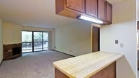 This single-floor unit is designed with an open-concept layout that connects the kitchen and living area into one cohesive space. The kitchen features rich, dark wood upper cabinetry paired with a light butcher block countertop breakfast bar, offering both functional workspace and a casual dining surface. Under-cabinet fluorescent lighting provides practical task illumination above the prep area.

The kitchen flows seamlessly into the carpeted living area, which benefits from natural light streaming through a full-width sliding glass door. The slider opens to a private balcony enclosed by a metal railing, with a backdrop of mature trees providing a sense of privacy and a natural outdoor view. The neutral-toned walls and light carpet throughout the living space create a clean, understated aesthetic. A doorway visible from the main living area leads to the remaining portions of the unit, suggesting additional rooms beyond the open-plan zone. Overall, the space balances functional kitchen design with a comfortable, light-filled living area and a pleasant connection to the outdoors.