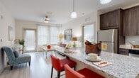 Located at 2770 Mystic Pointe Pkwy in Land O' Lakes, Florida, this single-floor residence offers a thoughtfully designed open-concept layout with a warm, coastal-inspired aesthetic throughout. The kitchen is a standout feature, showcasing rich dark wood cabinetry, stainless steel appliances, a classic white subway tile backsplash, and granite countertops that extend to a breakfast bar with seating — ideal for casual dining. Light wood-style flooring flows seamlessly into the living area, which is filled with natural light from large windows and a glass door leading to an outdoor access point. A ceiling fan completes the comfortable, airy atmosphere of the living room. The primary bedroom is generously sized with plush carpeting, a ceiling fan, and a large window dressed with elegant curtains. Its en-suite bathroom features a granite-topped vanity with dark wood cabinetry, tile flooring, and a glass-enclosed shower. A walk-in closet with built-in shelving provides ample storage and connects conveniently to the bathroom. A second bedroom offers a cozy retreat with carpeted floors and neutral tones. A full guest bathroom with a tub-shower combination completes the layout. Throughout, the space maintains a bright, neutral palette accented by coastal décor touches.