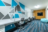 This dynamic single-floor workspace is designed to inspire productivity and collaboration through a carefully curated, energetic aesthetic. The space is anchored by a striking geometric mural rendered in teal, charcoal, and gray, creating a bold visual focal point along the main workstation wall. A long, continuous white desk runs the length of this wall, lined with ergonomic rolling chairs and clear acrylic privacy dividers that define individual workstations while maintaining an open, connected feel. Built-in drawer units beneath the desk provide convenient storage for each user. The flooring throughout features a patterned carpet tile in gray and teal tones, tying the color palette together cohesively. A dedicated conference area occupies one section of the space, furnished with a gray wood-tone table, bright yellow chairs, and a wall-mounted display screen set against a bold teal accent wall. An additional relaxed zone offers bean bag seating in front of a wall-mounted monitor on a warm mustard yellow accent wall, providing a more casual setting for collaboration or breaks. Mini-split HVAC units ensure climate control throughout. High clerestory windows along one wall allow natural light to filter in, complementing the overhead flush-mount lighting. The overall ambiance is modern, functional, and visually stimulating.