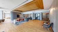 This single-floor space presents a contemporary lobby and amenity area defined by a refined blend of warm and neutral materials. The main lobby immediately draws attention with its striking slatted wood ceiling panels, recessed lighting, and light hardwood flooring that flows throughout the entire space, creating a sense of visual continuity. A central communal table with vibrant orange chairs anchors the open lounge area, positioned atop a patterned area rug that adds depth and texture to the floor plane. Steel-framed glass partitions divide portions of the space while maintaining visual openness, offering a glimpse into an adjacent meeting or dining area beyond. Near the entry vestibule, floor-to-ceiling glass doors and windows flood the transitional zone with natural light, where a compact round table and pendant globe fixtures create an inviting nook. A secondary seating area features a built-in L-shaped bench upholstered in warm caramel tones with textured gray walls and a sculptural wall sconce, lending a cozy, boutique atmosphere. A corridor extends toward a mailroom area, flanked by additional seating with paired lounge chairs against a textured feature wall. Throughout, the design strikes a balance between functional utility and sophisticated aesthetic, making this a welcoming and well-appointed communal environment.