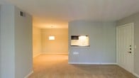This single-floor space showcases an open-concept layout that seamlessly connects the living and dining areas. The living area is finished with neutral-toned wall-to-wall carpeting, creating a warm and comfortable foundation throughout. Soft, light grey walls provide a clean, modern backdrop that complements the overall aesthetic.

A defining architectural feature of this space is the built-in pass-through kitchen window, illuminated by under-cabinet lighting that highlights dark cabinetry and a countertop with a sink. This functional design element offers a convenient connection between the kitchen and the main living area while maintaining a sense of visual openness.

The adjacent dining area is distinguished by a pendant light fixture suspended from the ceiling, casting warm, focused illumination over the space. The smooth white ceiling and consistent flooring throughout the open areas contribute to a cohesive and unified feel. Clean white baseboards frame the rooms neatly, adding a crisp finishing detail. The overall ambiance is calm and neutral, offering a versatile canvas well-suited for a variety of interior styles and furnishings.