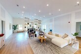 Located at Camelot Crossing in Valdosta, Georgia, this single-floor community space offers a generously sized, open-concept layout designed for both relaxation and recreation. The interior is anchored by warm light hardwood flooring that runs throughout, complemented by bright white walls and soaring ceilings that enhance the sense of openness and airiness.

The main living area is thoughtfully arranged with a plush sectional sofa, contemporary accent chairs, and a round wooden coffee table set atop a textured area rug — creating an inviting lounge zone. A large wall-mounted flat-screen television and a coordinating media console provide a dedicated entertainment focal point. A sleek pool table with a modern finish adds a recreational element, making the space ideal for socializing.

Recessed lighting and a striking globe pendant chandelier provide layered illumination, while French doors along one wall invite natural light and offer views to the exterior. A stylish open shelving unit serves as a partial room divider near a curved reception-style counter, adding both function and visual interest. A built-in beverage cooler and adjacent storage areas contribute practical amenities. The overall ambiance is clean, contemporary, and welcoming — well-suited for community gatherings and everyday leisure.