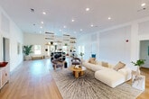 Located at Camelot Crossing in Valdosta, Georgia, this single-floor community space offers a generously sized, open-concept layout designed for both relaxation and recreation. The interior is anchored by warm light hardwood flooring that runs throughout, complemented by bright white walls and soaring ceilings that enhance the sense of openness and airiness.

The main living area is thoughtfully arranged with a plush sectional sofa, contemporary accent chairs, and a round wooden coffee table set atop a textured area rug — creating an inviting lounge zone. A large wall-mounted flat-screen television and a coordinating media console provide a dedicated entertainment focal point. A sleek pool table with a modern finish adds a recreational element, making the space ideal for socializing.

Recessed lighting and a striking globe pendant chandelier provide layered illumination, while French doors along one wall invite natural light and offer views to the exterior. A stylish open shelving unit serves as a partial room divider near a curved reception-style counter, adding both function and visual interest. A built-in beverage cooler and adjacent storage areas contribute practical amenities. The overall ambiance is clean, contemporary, and welcoming — well-suited for community gatherings and everyday leisure.