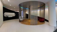 This striking single-floor space showcases a bold, contemporary design that blends futuristic aesthetics with luxurious comfort. The centerpiece is a large, circular enclosure defined by floor-to-ceiling shimmering metallic chain curtains that create a semi-transparent partition, offering both visual intrigue and a sense of intimate enclosure. Within this pod, warm-toned flooring contrasts beautifully with the sleek metallic ceiling above, which features a spectacular cascading crystal chandelier that serves as a breathtaking focal point. A vibrant blue LED strip runs along the inner perimeter of the circular ceiling, casting a cool, ambient glow that enhances the space's modern atmosphere. Inside the pod, a white sculptural egg chair and a deep red sectional sofa provide stylish seating options. Beyond the pod, the surrounding open floor area features large-format white tiles that amplify the sense of spaciousness and light. A textured dark feature wall with an organically shaped, illuminated oval mirror adds a dramatic design element to the perimeter of the space. The overall ambiance is sophisticated and avant-garde, combining dramatic lighting, curved architectural forms, and carefully curated furnishings to create an immersive and visually captivating environment.