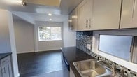 This single-floor space offers a thoughtfully designed open layout that seamlessly connects the kitchen to the living area. The kitchen is a standout feature, showcasing dark stone countertops that contrast elegantly with cream-toned upper cabinetry equipped with sleek bar-pull hardware. A striking mosaic tile backsplash in shades of blue, gray, and black adds a decorative focal point and visual texture to the space. A stainless steel double-basin sink is positioned beneath a frosted window, allowing natural light to filter in. Recessed lighting in the ceiling provides consistent, even illumination throughout. The kitchen flows naturally into an adjacent living area, which benefits from a large window that invites ample natural light, creating a bright and welcoming atmosphere. Dark hardwood-style flooring runs throughout, grounding the space with a warm, cohesive aesthetic that complements the modern finishes. Clean white walls and minimalist architectural details give the space a fresh, contemporary feel. Overall, this single-story layout is well-suited for those seeking a functional, stylish living environment with modern design elements and an open, connected floor plan.