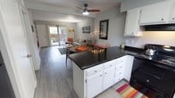 This single-floor residence offers a thoughtfully designed open-concept layout that connects the kitchen and living area in one cohesive, flowing space. The kitchen is appointed with crisp white cabinetry, dark countertops, and a peninsula that serves as both a prep surface and a natural divider between cooking and living zones. The kitchen seamlessly transitions into the living room, which is anchored by light-toned hardwood-style flooring that extends throughout the space, lending warmth and visual continuity. A ceiling fan with an integrated light fixture adds both function and a casual, modern touch to the living area. Natural light pours in through a sliding glass door at the far end of the living room, which opens directly onto a private outdoor patio, creating an inviting indoor-outdoor connection. The living area provides a comfortable and versatile gathering space with ample room for seating and décor. Neutral gray walls serve as a clean backdrop, allowing the space's architectural elements and finishes to take center stage. The overall ambiance is bright, airy, and welcoming, blending practical design with an approachable modern aesthetic suited for everyday living.