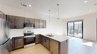 Located at 1301 Mulberry St in the heart of Des Moines, Iowa, this thoughtfully designed one-bedroom, one-bathroom unit offers a modern urban living experience. The open-concept kitchen and living area serves as the heart of the home, featuring rich dark-toned cabinetry, light quartz countertops, and a full suite of stainless steel appliances including a refrigerator, electric range, built-in microwave, and dishwasher. Elegant globe pendant lights with brass accents hang above the peninsula, adding a stylish touch. Light hardwood flooring flows throughout the main living areas, enhancing the bright and airy atmosphere created by large floor-to-ceiling windows and sliding glass doors that open to a private balcony with urban skyline views. An exposed metal ductwork detail adds an industrial accent to the space. The bedroom is carpeted for comfort, featuring a walk-in closet and its own access to the balcony through a sliding glass door, flooding the room with natural light. The bathroom is finished with a dark wood vanity, white countertop, modern globe light fixture, and a full soaking tub with shower. Clean white walls and recessed lighting throughout complete this contemporary, move-in ready urban retreat.