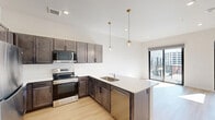 Located at 1301 Mulberry St in the heart of Des Moines, Iowa, this thoughtfully designed one-bedroom, one-bathroom unit offers a modern urban living experience. The open-concept kitchen and living area serves as the heart of the home, featuring rich dark-toned cabinetry, light quartz countertops, and a full suite of stainless steel appliances including a refrigerator, electric range, built-in microwave, and dishwasher. Elegant globe pendant lights with brass accents hang above the peninsula, adding a stylish touch. Light hardwood flooring flows throughout the main living areas, enhancing the bright and airy atmosphere created by large floor-to-ceiling windows and sliding glass doors that open to a private balcony with urban skyline views. An exposed metal ductwork detail adds an industrial accent to the space. The bedroom is carpeted for comfort, featuring a walk-in closet and its own access to the balcony through a sliding glass door, flooding the room with natural light. The bathroom is finished with a dark wood vanity, white countertop, modern globe light fixture, and a full soaking tub with shower. Clean white walls and recessed lighting throughout complete this contemporary, move-in ready urban retreat.