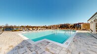 Located at 5315 Waters Edge Village Drive in Charlotte, NC, this outdoor amenity space is a standout feature of the Waters Edge Village community. At its centerpiece is a generously sized, resort-style swimming pool with crystal-clear turquoise water, surrounded by a broad stone-paver deck finished in a classic travertine style. The pool deck is lined with an array of chaise lounges, providing ample space for sunbathing and relaxation under the open Carolina sky.

Adjacent to the pool is a charming covered cabana structure with white columns and decorative lantern lighting, offering shaded seating and a welcoming retreat from the sun. A separate outdoor lounge patio features wicker furniture, a round drum coffee table, and lush landscaping, creating an intimate gathering space ideal for socializing. Multi-level covered balconies on the adjoining clubhouse building overlook the entire pool area, accented with ceiling fans for comfort.

Architectural details throughout the space reflect a refined, coastal-inspired aesthetic with clean white facades, arched windows, and latticed pergola structures that add visual interest and privacy. The surrounding community buildings frame the space, while open sightlines to the sky and mature treeline give the area a bright, airy ambiance throughout the day.